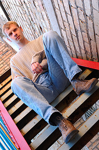 年轻时尚的人坐在砖墙附近的楼梯上浅黄色橙子毛衣男人脚步头发蓝色扶手鞋类牛仔裤图片