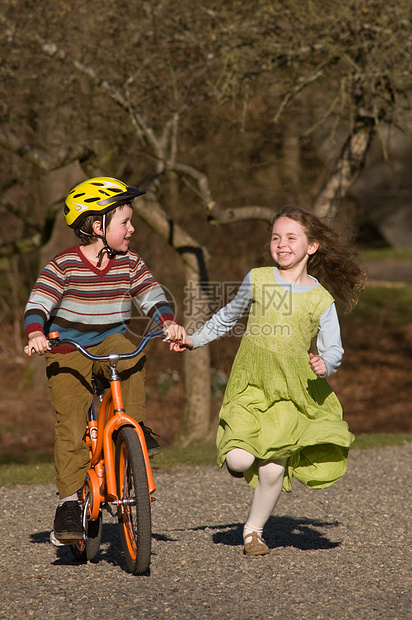男孩骑自行车 女孩跑在他身边兄弟姐妹练习头盔骑术兄弟姐姐绿色公园跑步毛衣图片