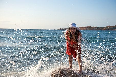 美丽的女孩在海浪中飘荡裙子日落海滩太阳享受热带乐趣假期飞溅岩石图片