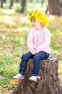 美丽的小姑娘 坐在石板上 坐着一朵满是青梅树叶的花圈里快乐压痛叶子女儿微笑季节孩子公园童年树桩图片