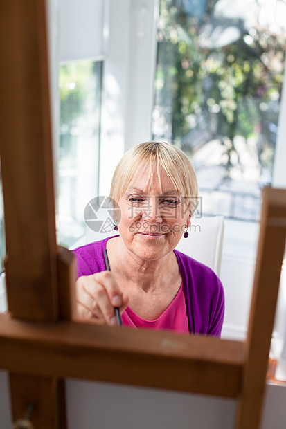 快乐的年长妇女在家里为取乐而绘画刷子画架老年热情艺术品画笔帆布奶奶调色板闲暇图片