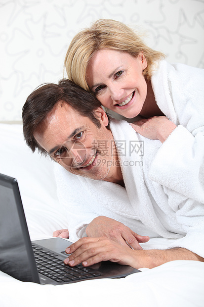 穿睡衣时在笔记本电脑上快乐的一对情侣个人浴衣微笑女士白色金发丈夫敷料男性妻子图片