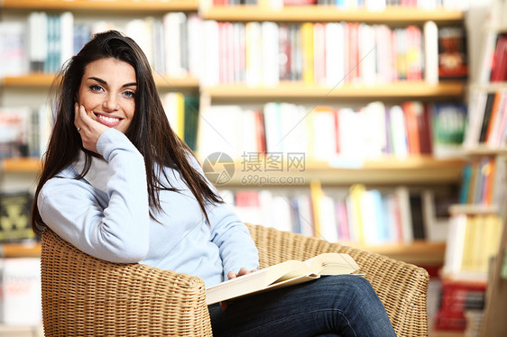 带着书在手的笑着微笑的女学生坐在椅子上学习书店青年店铺女孩科学书架女士教育书柜图片