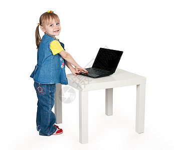 穿着蓝色牛仔裤的小女孩站在一张带笔记本电脑的桌子上图片