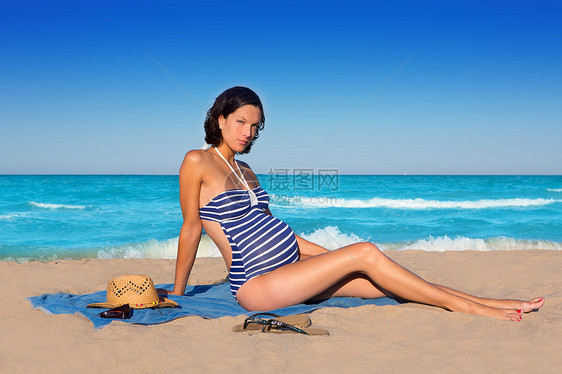 坐在蓝沙沙滩沙滩上的美丽的孕妇海洋蓝色支撑怀孕海滩泳装父母成人幸福假期图片