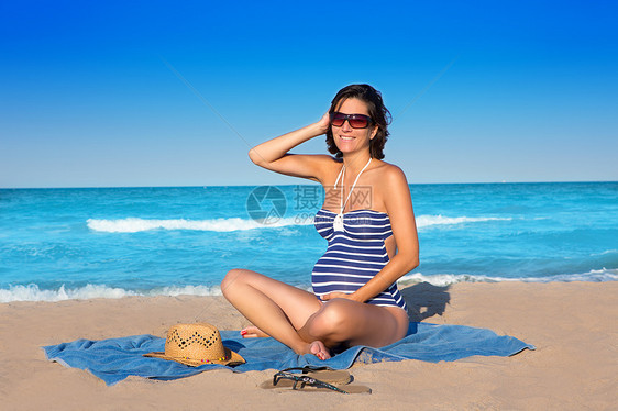 坐在蓝沙沙滩沙滩上的美丽的孕妇婴儿家庭泳装海滨假期女孩蓝色父母幸福母性图片