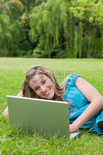 年轻微笑的小女孩在躺在草地上时使用笔记本电脑图片