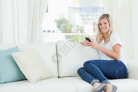 一个女人坐在沙发上 一边用她的手机 一边坐着金发金发女郎成人快乐划线长椅外表微笑电话女性图片