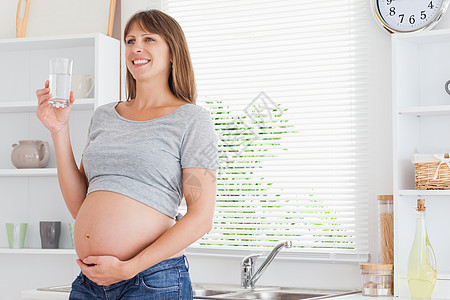 怀孕的孕妇在站立时拿着一杯水喝着微笑灰色女性冒充厨房亮度头发花朵女士窗户图片
