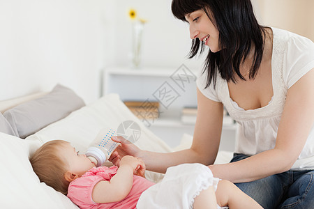 迷人的黑褐色女性在床上用瓶子喂婴儿说谎女孩儿子喜悦女儿金发乐趣生活幸福妈妈图片