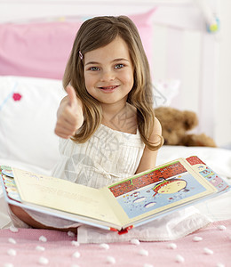 快乐的小女孩在床上看书时举起拇指背景图片