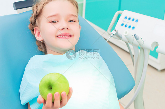 坐在牙医椅子上的女孩展示了一个绿苹果幸福外科卫生从业者工具蓝色工作矫正孩子女性图片