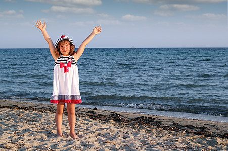 快乐的小女孩 在海滩上举起手帽子假期白色闲暇喜悦海浪微笑海滨蓝色裙子图片