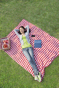 微笑着的年轻女人躺在背上 手抱在头后 坐在毯子上 在公园里放松图片