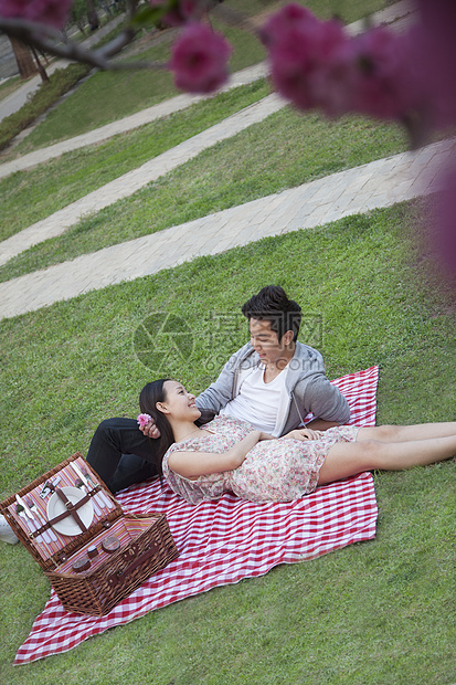 在公园野餐时 一对幸福的年轻夫妇一起躺在毯子上 前景是鲜花图片