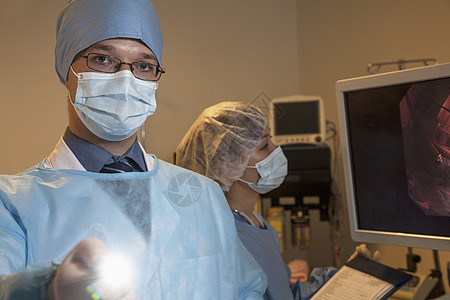 年轻外科医生的肖像 他用医疗仪器瞄准照相机 照亮光检查年轻人面具器材口罩手术光束手术帽防护内窥镜图片