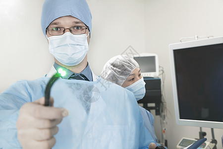 年轻外科医生的肖像 他用医疗仪器瞄准照相机 照亮光职业医护人员年轻人手术室权威眼镜团队面具摄影工作服图片