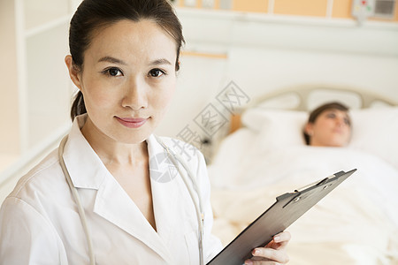 微笑的医生拿着病历 背景是病人躺在医院病床上的肖像棕色疾病两个人记录女性病房医疗快乐医护人员监视图片