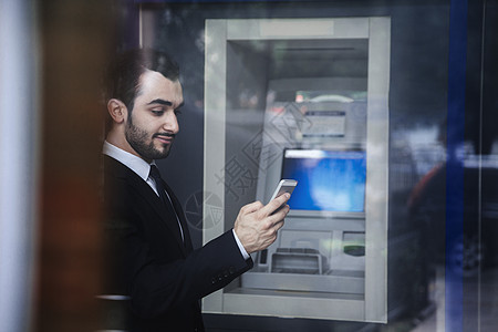 站在自动取款机前看着手机的年轻商务人士笑着看电话头发外观柜员机玻璃胡子微笑收腰胡须摄影反射图片