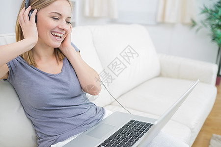 快乐的妇女通过笔记本电脑耳机听音乐娱乐沙发长椅家庭生活房子金发女性头发技术音乐图片