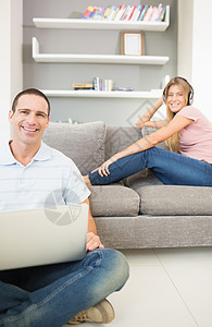 坐在地板上的男人用笔记本电脑与女人一起在沙发上听音乐 两人都对着镜头微笑图片