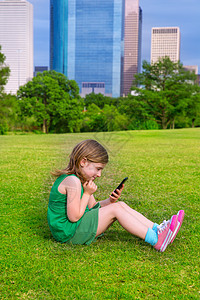 坐在C区公园草坪上玩智能手机的Blond女孩孩子金发办公室地标蓝色技术建筑物景观草地电话图片