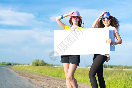 两名年轻女性站在空白的横幅上站着旅行便车纸板游客自由路线女士天空成人假期图片