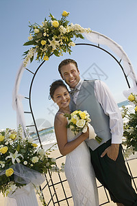 海滩大拱门下肖像的新娘和Groom海洋关系结婚正装表情连衣裙婚礼服风格玫瑰新郎背景图片