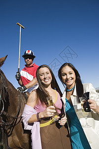 快乐的女朋友和马球手一起喝香槟的肖像 在背景中坐在马背上图片