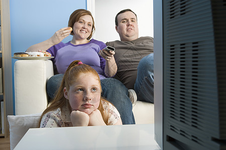 超重家庭在沙发上看电视专注饮食肥胖父亲电视中年牛仔裤休闲小吃女性图片
