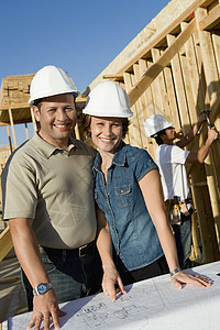 一对多族裔夫妇在硬达帽的肖像 建筑工地有蓝图图片