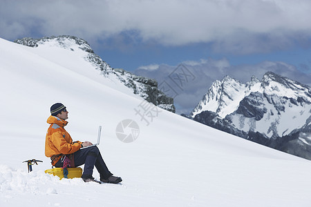 在雪山坡上使用笔记本电脑的男性徒步者的侧边视图图片