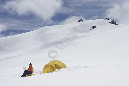 使用膝上型电脑在雪山坡上搭帐篷的男性徒步旅行者的侧面视图勘探旅行笔记本上网高度技术远程顶峰远足者娱乐图片
