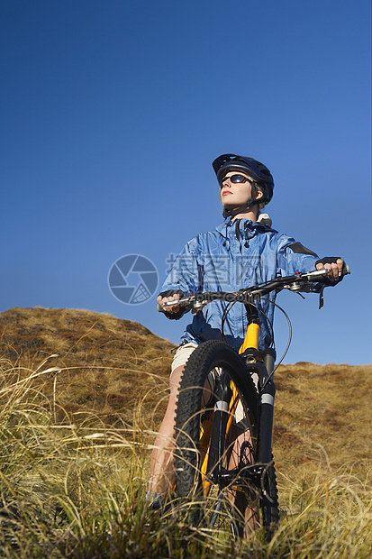 年轻女子坐在田地上骑自行车对抗清蓝天空国家安全骑术娱乐农村运输运动闲暇运动员爱好图片