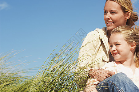 快乐的母亲和小女儿坐在长草地上 与蓝天对立图片