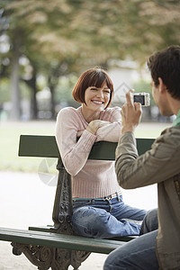 在公园长凳上拍摄微笑着的年轻女子的照片拍照娱乐刘海闲暇旅游深色边缘女士乐趣成人图片