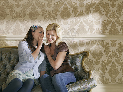 年轻女人对着墙纸 在沙发上向快乐朋友的耳朵低语图片