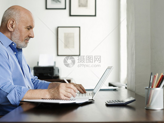 使用笔记本电脑和在主桌上写笔记本的成熟男子侧面视图图片