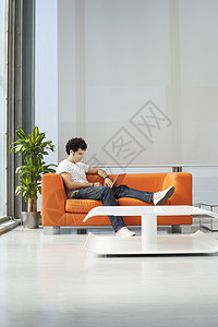 在办公室接待室的橙色沙发上使用笔记本电脑的一名年轻人全身图片
