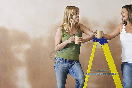 两个年轻女人靠在脚梯上 与杯子对着棕墙图片