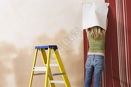看着一个金发的 年轻女孩上挂壁纸背心成人女性牛仔布创造力公寓维修房间墙纸梯子图片