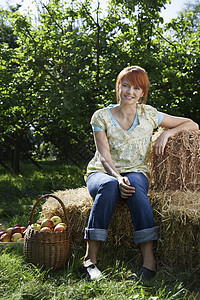 一位坐在果园附近的干草篮上的年轻女子的全长肖像干草农村食物女性闲暇篮子稻草牛仔裤植物树木图片
