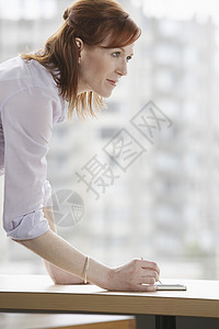 一位在办公桌前写字的红头发女商务人士的侧面视图背景图片