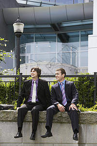 坐在办公室外墙上的两名青年商务人士全身在办公室外背景图片