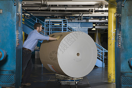 一个男人在报纸工厂里 推着一大卷纸的侧面景象图片