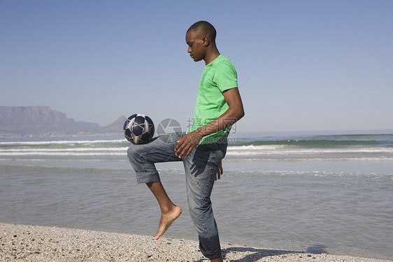 一位年轻非裔美国人在海滩踢足球的侧面观图片