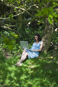 在树林中使用笔记本电脑的孕妇女子蓝色公共公园头发林地树干农村赤脚女士技术图片