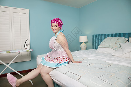 一位快乐的年轻女士坐在床上 背着熨衣板在背景中被烫衣服的肖像背景图片