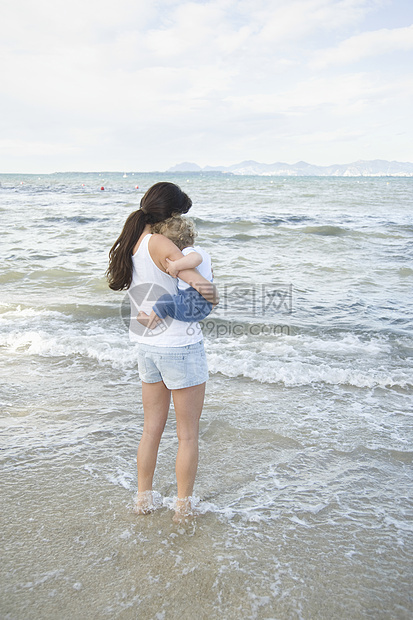 一位女士拿着一只脚在浅水中的幼儿 站在海正前方 背着远山的山峰背景图片