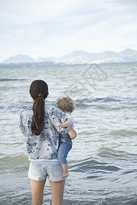 一位女士拿着一个幼儿手站在海面前 有着远处的山峰背景图片